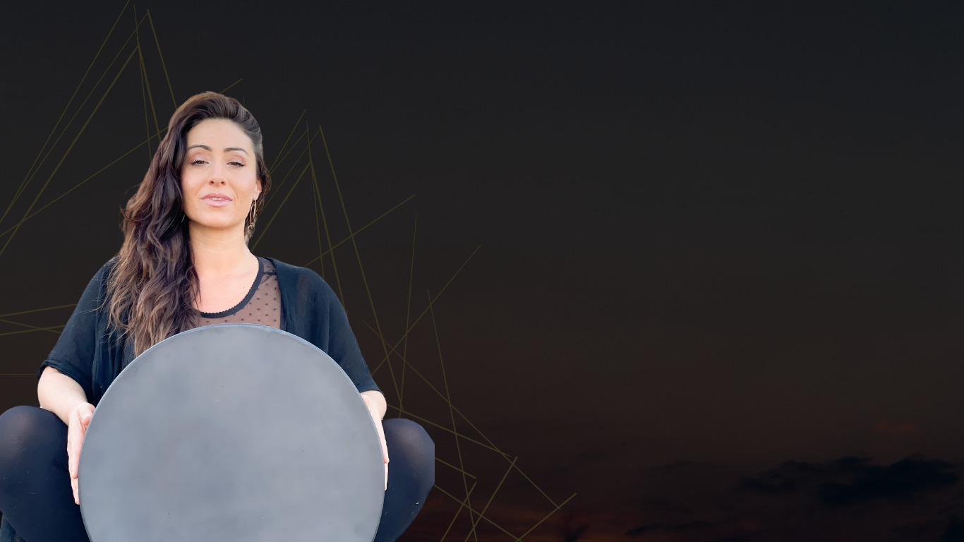 Larisa seated with her frame drum against a dusk sky, holding space for the circle