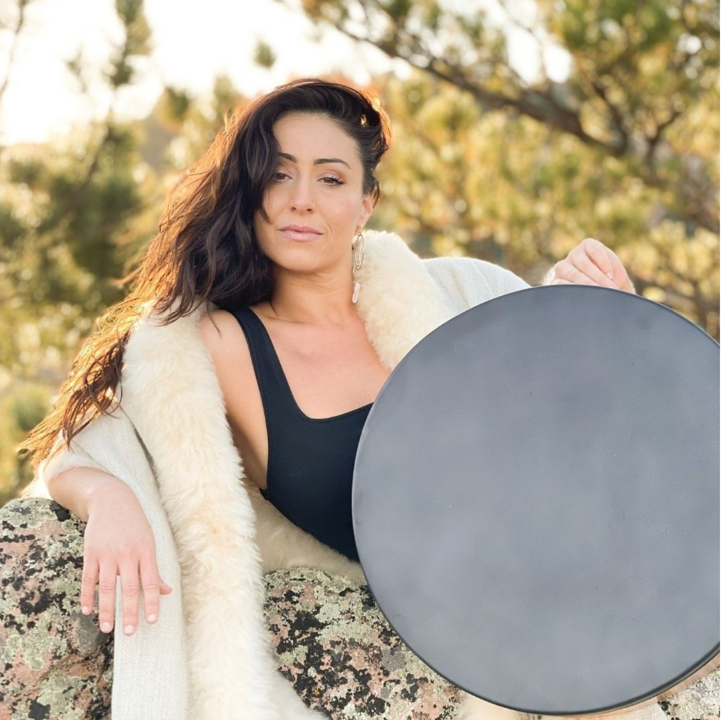Larisa, creator of the Vocal Resonance Method, seated outdoors in golden light holding her frame drum